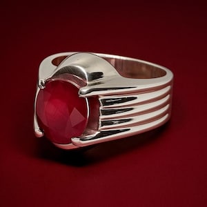 May include: A silver ring featuring a large, oval-cut, red gemstone. The ring has a wide band with horizontal grooves and a setting that securely holds the gemstone. The background is a deep red.