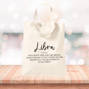 Könnte beinhalten: Eine cremefarbene Tragetasche mit dem Wort "Libra" in einem stilvollen Schriftzug. Unter dem Wort befindet sich eine Definition von Waage, die die Eigenschaften des Sternzeichens beschreibt. Die Tasche hat ein schlichtes Design mit einem Griff.