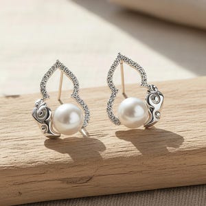 May include: A pair of silver earrings, each featuring a large, round pearl. The earrings have an openwork design with small, clear stones and scrollwork. They are displayed on a wooden surface. The pearls are the focal point.