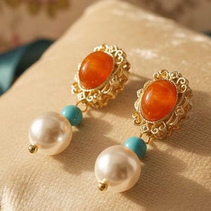 Puede incluir: Un par de pendientes dorados con piedras centrales ovaladas de color naranja. Cada pendiente presenta una gota de perla y una pequeña cuenta turquesa. Los pendientes se exhiben sobre una superficie de terciopelo beige.