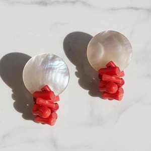 Pode incluir: Um par de brincos com um disco redondo de madrepérola iridescente e um aglomerado de coral vermelho vibrante. Os brincos são apresentados sobre uma superfície de mármore branco, com sombras que adicionam profundidade.