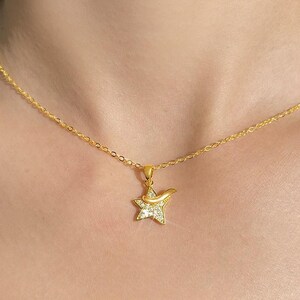 May include: A gold-colored necklace with a star pendant. The star pendant is encrusted with small, clear gemstones and has a curved design element. The chain is delicate and also gold-colored. The necklace is displayed against a skin-toned background.