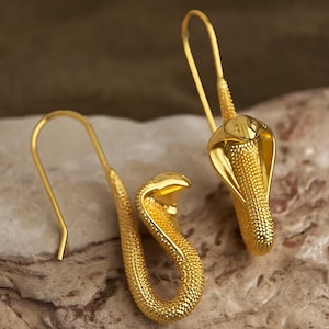 Peut inclure: Une paire de boucles d'oreilles cobra dorées. Les boucles d'oreilles présentent un corps texturé et un capuchon évasé, avec une fermeture à crochet. Le design est détaillé et les boucles d'oreilles sont présentées sur une surface aux tons neutres.