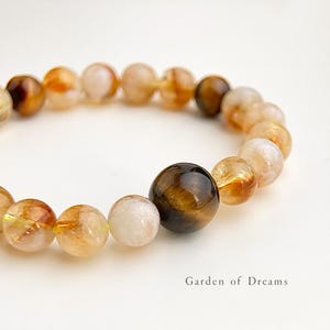 May include: A beaded bracelet featuring a variety of polished gemstone beads in shades of brown, yellow, and white. The bracelet has a mix of opaque and translucent beads, with a large, dark brown bead as a focal point. The text "Garden of Dreams" is visible.