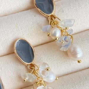 May include: Gold-toned dangle earrings. Each earring features a dark gray oval-shaped top, connected to a cluster of white and iridescent stones, and a single pearl. The earrings are displayed on a cream-colored surface.