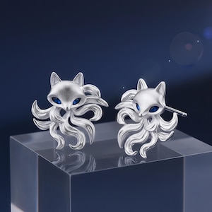 May include: A pair of silver-colored fox earrings. Each earring features a fox head with blue eyes and a textured body, surrounded by flowing, stylized fur. The earrings are displayed on a clear, rectangular stand against a dark blue background.