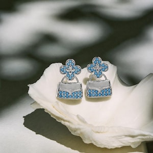 Könnte beinhalten: Ein Paar silberfarbene Ohrringe in Form von Miniatur-Handtaschen. Jeder Ohrring zeigt oben ein vierblättriges Kleeblatt-Motiv, das mit kleinen, runden, leuchtend blauen Steinen besetzt ist. Der Boden der Handtasche ist ebenfalls mit blauen Steinen verziert.