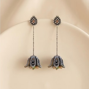 May include: A pair of long drop earrings featuring a teardrop-shaped stud with a central peach-colored stone surrounded by small white beads. A thin metal chain connects the stud to a bell-shaped pendant with blue, grey, and green accents.
