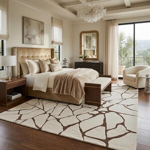 May include: A large area rug featuring an off-white background with a dark brown, irregular, stone-like geometric pattern. The rug is positioned in a bedroom with a tufted beige headboard, a wooden bench, and a cream-coloured armchair nearby.