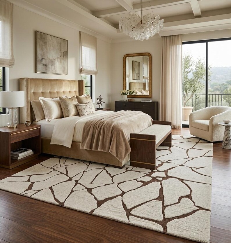 May include: A large area rug featuring an off-white background with a dark brown, irregular, stone-like geometric pattern. The rug is positioned in a bedroom with a tufted beige headboard, a wooden bench, and a cream-coloured armchair nearby.