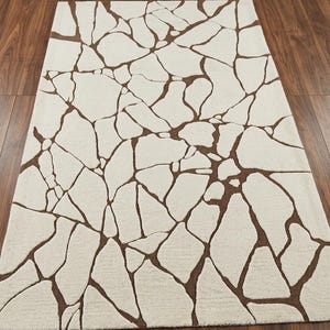 ​Rectangular handtufted wool area rug featuring an organic cracked earth mosaic pattern in earthy brown on a cream base for modern minimalist decor.