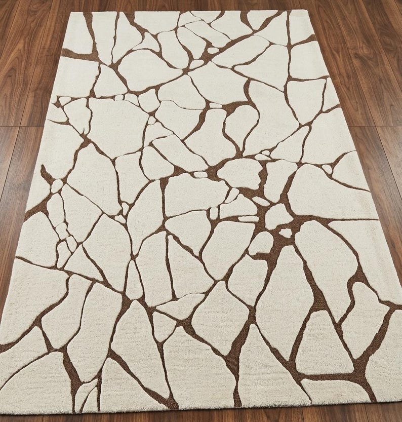 ​Rectangular handtufted wool area rug featuring an organic cracked earth mosaic pattern in earthy brown on a cream base for modern minimalist decor.