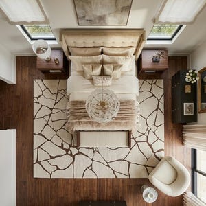 May include: A top-down view of a bedroom featuring a large bed with a tufted beige headboard, layered cream bedding, and a decorative throw. A rug with a dark brown cracked stone pattern covers the dark wood floor, alongside a round chair.