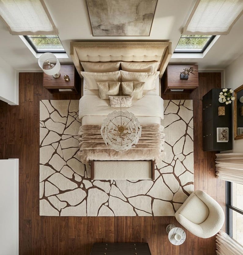 May include: A top-down view of a bedroom featuring a large bed with a tufted beige headboard, layered cream bedding, and a decorative throw. A rug with a dark brown cracked stone pattern covers the dark wood floor, alongside a round chair.