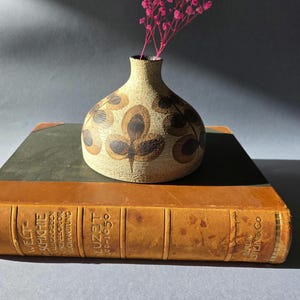 Könnte beinhalten: Eine kleine, beige Keramikvase mit braunen blattartigen Mustern, die einen Zweig rosa Blüten enthält. Die Vase steht auf einem Vintage-Buch mit braunem Ledereinband und goldfarbenen Buchstaben. Der Hintergrund ist ein neutrales Grau.