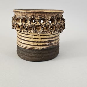 Könnte beinhalten: Eine handgefertigte Keramikvase mit einzigartigem Design. Die Vase hat einen dunkelbraunen Sockel, einen mittelbraun gestreiften Mittelteil und eine strukturierte Oberseite mit kreisförmigen Öffnungen. Die Vase ist etwa 10 cm hoch.
