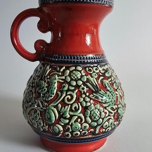 Pode incluir: Jarro de cerâmica vermelho com alça e corpo bulboso. A parte superior e a base são vermelhas, com uma faixa azul ao redor do pescoço e da base. O corpo apresenta um desenho floral e de pássaros verdes e brancos.