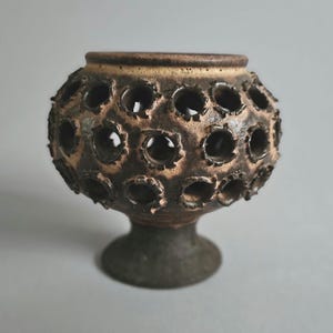 Könnte beinhalten: Eine braune Keramikvase mit einzigartigem Design. Die Vase hat einen runden Körper mit zahlreichen kreisförmigen Öffnungen, einen hellbraunen Rand und einen dunkelbraunen Sockel. Die Textur wirkt rau und handgefertigt.