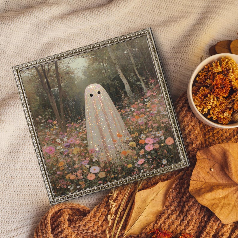 May include: A framed art print featuring a whimsical ghost in a field of colorful flowers. The ghost is white with black eyes and glitter accents. The frame is silver with a decorative border. A bowl of dried flowers and a fall leaf are also visible.