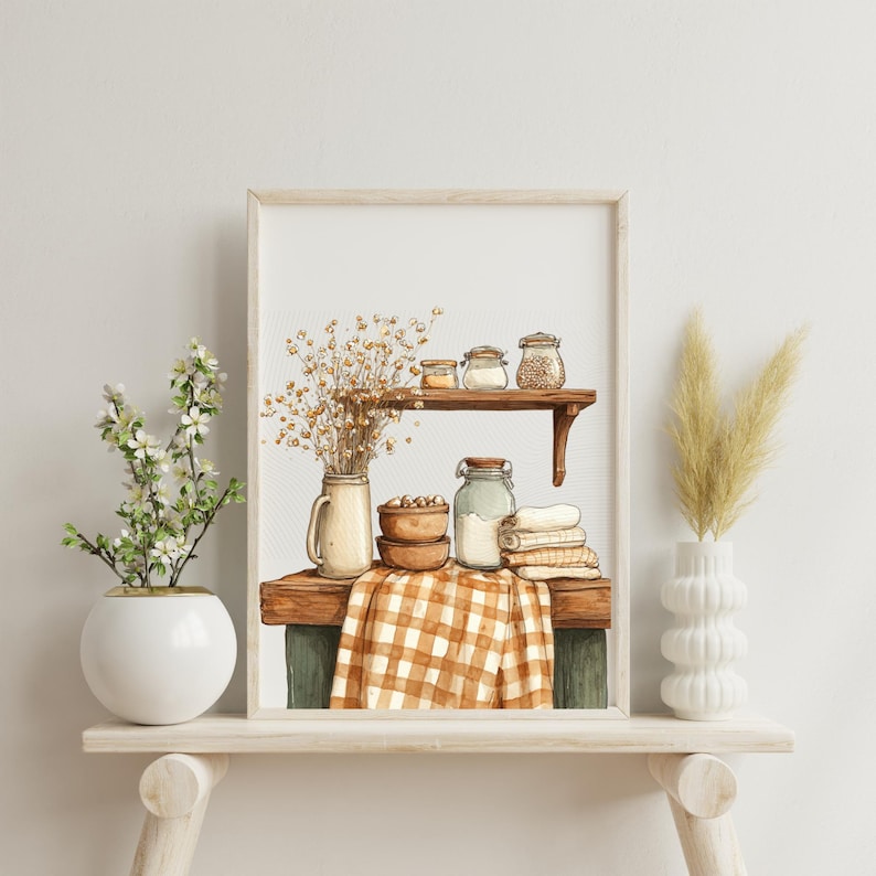 May include: Watercolor print of a rustic kitchen scene. A wooden shelf displays jars, bowls, and a checkered cloth. Dried flowers and decorative vases flank the framed artwork, creating a cozy, farmhouse aesthetic.