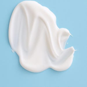 May include: A smooth, white cream product, possibly a lotion or moisturizer, is swirled on a light blue background. The cream has a glossy appearance and a thick consistency, with a few pointed edges.