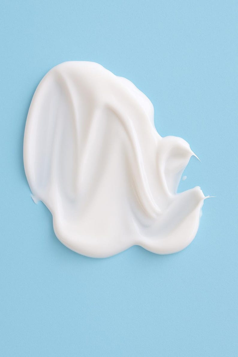 May include: A smooth, white cream product, possibly a lotion or moisturizer, is swirled on a light blue background. The cream has a glossy appearance and a thick consistency, with a few pointed edges.