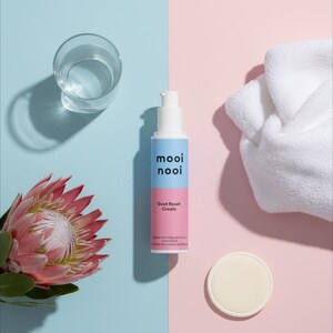 May include: A skincare product display featuring a white and blue bottle of "mooi nooi Quiet Reset Cream", a glass of water, a white towel, a round pad, and a pink protea flower. The background is split between light blue and pink.
