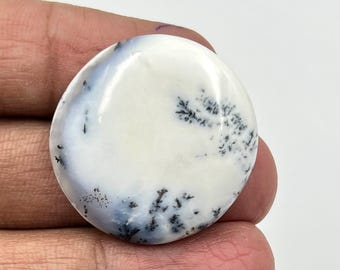 May include: A round, polished gemstone with a white base and intricate black and gray dendritic inclusions. The stone has a smooth, glossy surface and is held in a hand. The gemstone is a natural, unique piece.