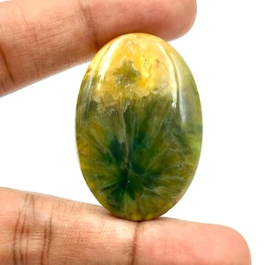 May include: An oval gemstone displaying a mix of yellow and green shades. The stone has a distinctive pattern, reminiscent of a burst of light or a floral motif. The polished surface reflects light, enhancing the natural colours and textures.