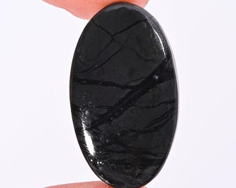 Puede incluir: Una piedra preciosa negra pulida, de forma ovalada, con vetas naturales. La piedra se sostiene entre dos dedos, mostrando su superficie lisa y reflectante. El color oscuro y los patrones únicos sugieren su uso en joyería o como elemento decorativo.