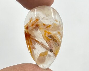 May include: A clear, teardrop-shaped gemstone with orange and tan mineral inclusions. The polished stone is held between fingers, highlighting its translucent quality and natural patterns. A unique piece of jewellery.