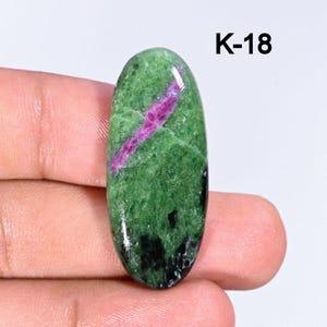 May include: An oval gemstone, predominantly green with a striking pink stripe. Dark black patches are visible. The text "K-18" is in the upper right corner. The stone is held in a hand.