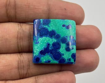 May include: A square-shaped gemstone with a polished surface. The stone features a vibrant mix of teal and deep blue hues, with a speckled pattern. The gemstone is held in a hand, showcasing its size and color variations.