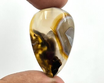 May include: A polished, teardrop-shaped gemstone. The stone displays a blend of light brown, dark brown, and white hues, accented with yellow. The gemstone is held between two fingers.