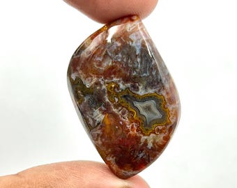 May include: A polished gemstone with a teardrop shape. The stone features a mix of colours, including shades of red, brown, white, and gold. The pattern has a central eye-like design with concentric rings. The gemstone is held between two fingers.