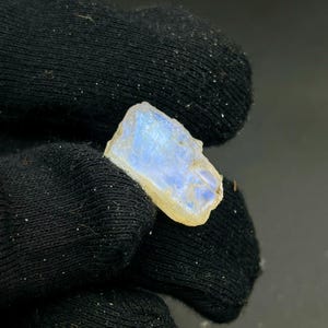 May include: A raw, uncut moonstone gemstone with a pale blue and yellow hue. The stone is held against a black background, showcasing its natural, irregular shape and subtle iridescence. The moonstone appears to be a small, rough-hewn crystal.