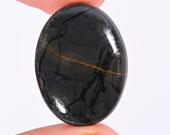 Puede incluir: Una gema ovalada pulida, de color oscuro, casi negro. Finas vetas claras y una fina línea marrón dorada recorren la superficie. La piedra se sostiene entre los dedos, resaltando su acabado liso y reflectante. Ideal para joyería.