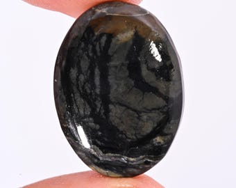 Puede incluir: Una piedra preciosa de forma ovalada con una superficie pulida. La piedra presenta un patrón oscuro y arremolinado de negro y gris, con toques de marrón en la parte superior. La piedra preciosa se sostiene entre dos dedos, mostrando su tamaño y forma.