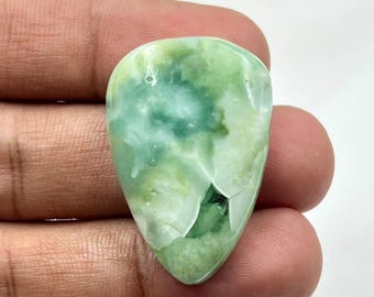May include: A polished, teardrop-shaped gemstone with a mix of light and dark green hues. The stone has a marbled appearance with areas of opaque white and translucent green. The gemstone is held in a hand against a white background.