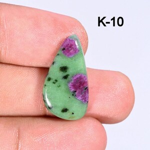 Peut inclure: Une pierre précieuse polie avec une base vert clair, présentant des inclusions magenta vives et vert foncé. La pierre est en forme de goutte d'eau et tenue dans une main. Le texte "K-10" est imprimé en noir.