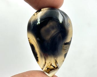 May include: A polished, teardrop-shaped gemstone with a striking pattern of black, tan, and cream swirls. The stone is held between fingers, showcasing its smooth, glossy surface and natural colour variations. The gemstone is a unique piece.