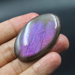 Puede incluir: Gema de labradorita de forma ovalada con superficie pulida. La piedra muestra un vibrante juego de colores, con tonos morados y toques dorados. La gema se sostiene en una mano, mostrando su tamaño y forma. La piedra mide aproximadamente 5 cm de largo.