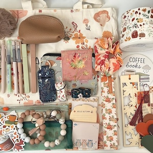 May include: A collection of autumn-themed stationery and accessories including a canvas tote bag, a ceramic mug with pumpkin and leaf patterns, pens, a brown pouch, a scrunchie, bookmarks, stickers, sticky notes, and a beaded keychain with a tassel.