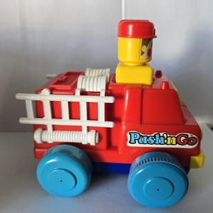 May include: A red plastic toy fire truck with blue wheels and a white ladder. The toy has a yellow and red figure on top and the words "Push'n Go" on the side.