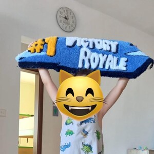 May include: A blue and yellow rug with the words "VICTORY ROYALE" in white. The rug is held up by a person. The rug has a blue border and a yellow number one.