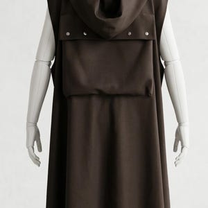 May include: A dark brown sleeveless hooded cloak with a large pocket and silver-colored snap closures. The cloak is displayed on a mannequin. The garment has a long, flowing design, suitable for cosplay or costume events.