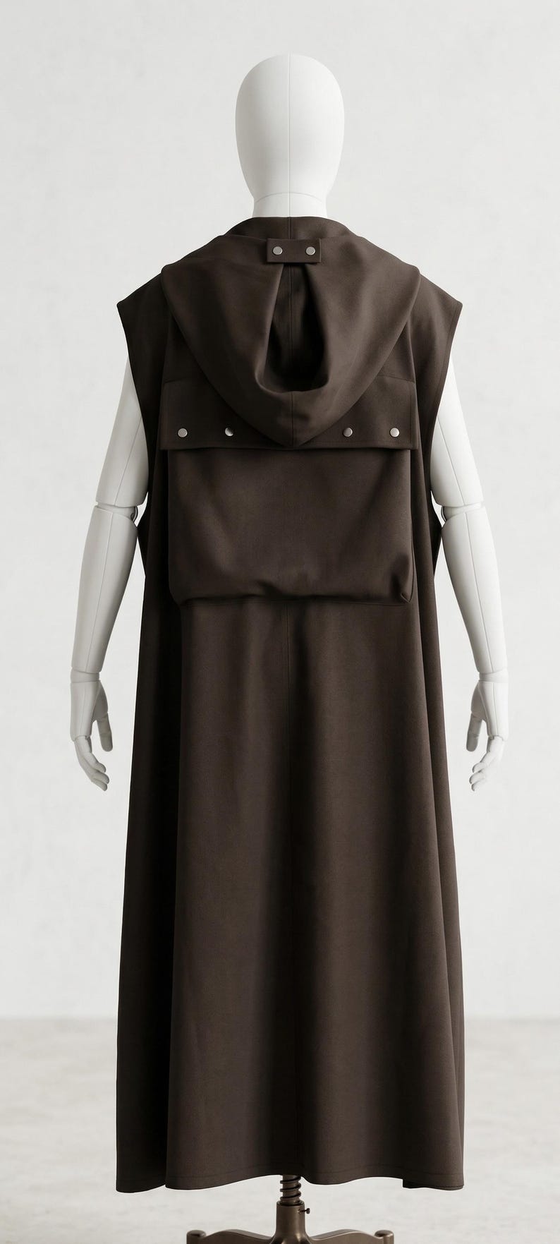 May include: A dark brown sleeveless hooded cloak with a large pocket and silver-colored snap closures. The cloak is displayed on a mannequin. The garment has a long, flowing design, suitable for cosplay or costume events.