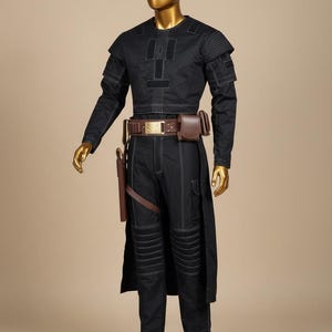 May include: Black costume with white stitching, featuring a long-sleeved top, pants, and a long coat. The outfit includes a brown belt with a gold buckle, a holster, and a pouch. The mannequin has a gold head and hands.