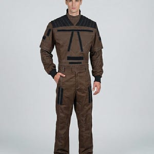 Puede incluir: Un mono marrón con detalles negros en los hombros, el pecho y los bolsillos. El mono tiene un cuello alto y mangas largas con puños negros. La persona lleva zapatos negros y tiene una mano en el bolsillo.