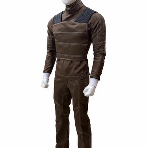 May include: Brown jumpsuit with white stitching details, featuring a high collar and shoulder straps. The long-sleeved top has white cuffs, and the pants have reinforced knee panels. This outfit is displayed on a mannequin.